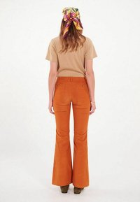 Pantalons en velours côtelé orange évasés avec poches arrière ; associés à une chemise beige à manches courtes et à un foulard à motif coloré noué dans les cheveux.