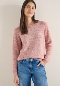 Femme aux longs cheveux bruns portant un pull en tricot rouge et blanc et un jean bleu, debout devant un mur beige clair uni.