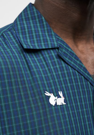 Πουκάμισο με πράσινο και ναυτικό καρό μοτίβο και γιακά με χαραγμένο άνοιγμα· διαθέτει λευκά κεντημένα κουνελάκια με κόκκινες λεπτομέρειες κοντά στο στήθος. Μαλακή υφή υφάσματος.