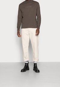 Långärmad, olivgrön bomullsskjorta med präglad logotyp, krämfärgade byxor med uppvikta ben och svarta läderankelboots med vita strumpor.