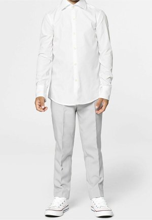 Barn stående iført en hvid skjorte med knapper, lysegrå bukser og hvide sneakers, mod en ensfarvet lys baggrund.