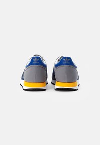 Dvojice šedých a modrých tenisek Adidas se žlutou a bílou podrážkou, zobrazené zezadu na čistém bílém pozadí.