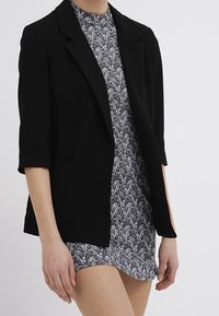 Černý blazer s třičtvrtinovými rukávy, který se vyznačuje strukturovaným designem a předními kapsami, nasazený na vzorované šaty v černé a bílé.