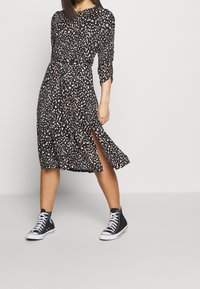 Frau, die ein schwarzes gepunktetes Midikleid mit einem seitlichen Schlitz und schwarzen High-Top-Sneakern trägt, steht vor einem schlichten, hellen Hintergrund.