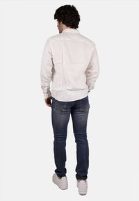 Camicia bianca a maniche lunghe con colletto a bottoni, abbinata a jeans in denim blu e sneakers bianche, vista da dietro.