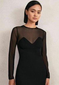 Tânără cu păr drept și negru, purtând o rochie lungă cu mâneci lungi, de culoare neagră, cu un top subțire și un corset cu dungi, stând în fața unui perete simplu de culoare deschisă.