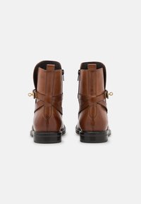 Bottines en cuir marron avec une finition lisse, dotées de fermetures éclair à l'arrière et de bandoulières décoratives. Semelles en caoutchouc noir avec une légère texture.