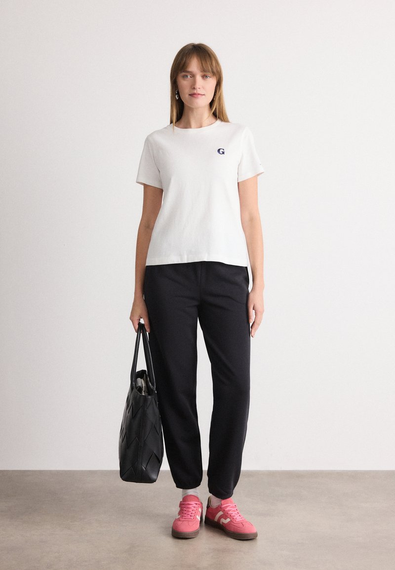 Weißes Baumwoll-T-Shirt mit kleinem navy Logo, kombiniert mit schwarzen Hosen. Eine schwarze Tragetasche und rosa Sneakers runden das Outfit ab.