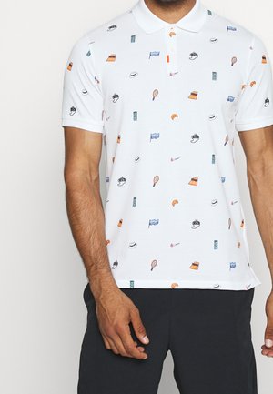 Polo blanc avec de petites icônes colorées comprenant des chapeaux, des raquettes de tennis, des croissants, des appareils photo, des drapeaux et des feux de signalisation, porté avec un pantalon noir.
