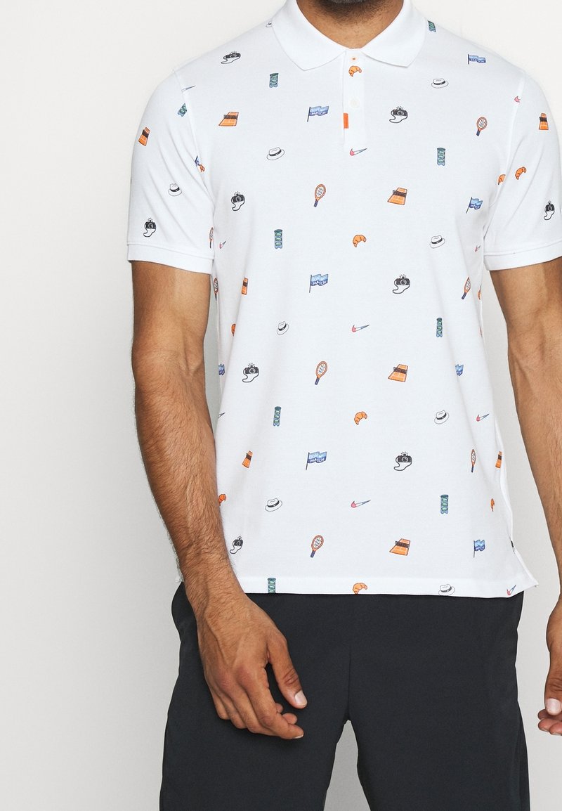 Polo blanc avec de petites icônes colorées comprenant des chapeaux, des raquettes de tennis, des croissants, des appareils photo, des drapeaux et des feux de signalisation, porté avec un pantalon noir.
