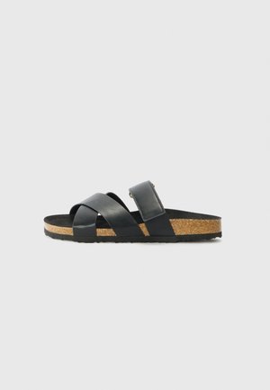 Sort sandal med korksål, to krydsede læderremme foran og en justerbar rem ved anklen på en grå baggrund.