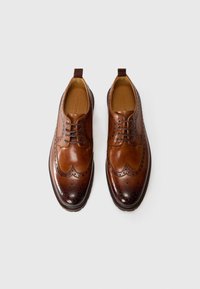 Brogues en cuir marron avec des perforations détaillées, un design courbé et une semelle superposée. Comprend un onglet décoratif au talon.
