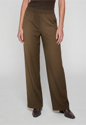 Personne portant un pantalon marron ample avec une ceinture élastique et des bottes marron foncé à bout pointu, debout devant un fond uni.