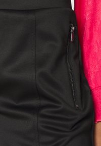 Schwarzer Skort aus elastischem Stoff, mit einer seitlichen Reißverschlusstasche und Falten-Design. Die Textur ist glatt und hat eine figurbetonte Silhouette.