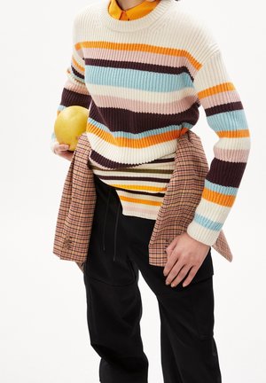 Persoon met een multikleurige gestreepte trui en een gelaagde geruite jas, die een geel stuk fruit vasthoudt, gekleed in zwarte broek tegen een witte achtergrond.