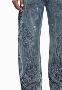 Τζιν παντελόνι από μπλε denim με φθαρμένη εμφάνιση, κεντημένα σχέδια chevron, ασημένια καρφιά και φθαρμένα άκρα. Χαλαρή εφαρμογή με διάφορες αποχρώσεις του μπλε.