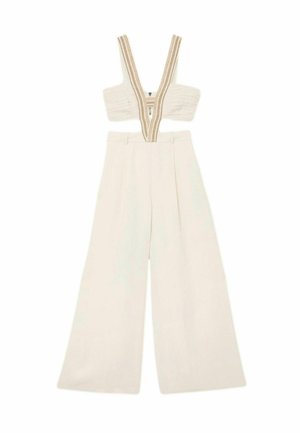 Naine punutud juustega, kellel on seljas beež värvi varrukateta lühike topp lõigatud detailidega ja kõrge vöökohaga laiade säärtega püksid valge seina taustal.