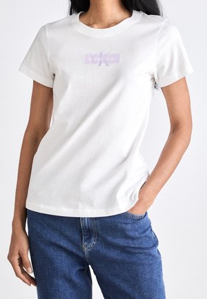 Kobieta ubrana w białą koszulkę Calvin Klein Jeans i niebieskie dżinsy, z jedną ręką w kieszeni, stojąca na jednolitym tle.