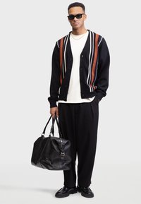 Schwarze Strickjacke mit orange- und weißen Streifen, weißes T-Shirt, weite schwarze Hosen, schwarze Leder-Duffel-Tasche, Sonnenbrille und schwarze Schuhe.