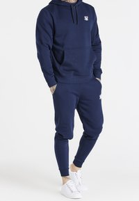 Navy blue hoodie with a front pocket and white logo, paired with matching joggers. Fabric appears smooth, fitted at cuffs and ankles.