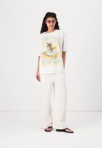 T-shirt grafica bianca con design di palma, abbinata a pantaloni bianchi larghi e testurizzati e infradito nere. Occhiali da sole semplici completano il look.