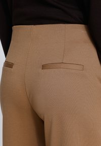 Gros plan d’une personne portant un pantalon marron taille haute avec deux poches passepoilées à l’arrière et un haut noir.