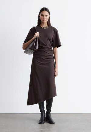 Kvinde iført en mørkebrun asymmetrisk kjole, sorte knælange støvler, guldkædehalskæde, der holder en matchende mørk håndtaske på skulderen.