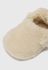 Crème-kleurige slipper gemaakt van zacht, gestructureerd nepbontmateriaal. Heeft een ronde neus en een comfortabele, gesloten ontwerp.