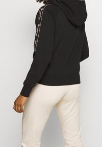 Felpa nera con cappuccio e accenti logo rosa lungo le maniche, tessuto morbido e design aderente. Abbinata a pantaloni chiari.