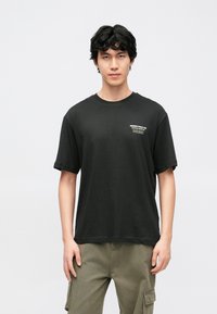 T-shirt de algodão preta com gola redonda, mangas curtas e um pequeno logótipo impresso no peito esquerdo. Combinada com calças cargo verde-oliva.