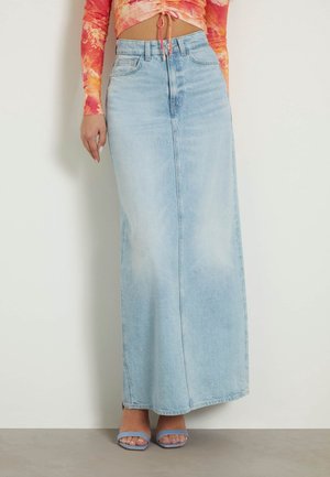 Femme portant une jupe maxi en denim bleu clair, un top court tie-dye orange et rouge, et des sandales ouvertes bleu clair, debout contre un mur blanc uni.