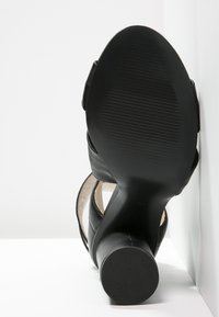 Musta korkokenkäsandaali pyöreällä korolla, leveillä remmeillä ja teksturoidulla pohjalla, näkymä pohjasta valkoista taustaa vasten.