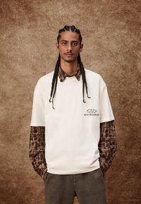 Kremaste majice s kariranim košuljom ispod koja sadrži smeđe i crne karirane uzorke; logo na grudima; ležeran kroj.