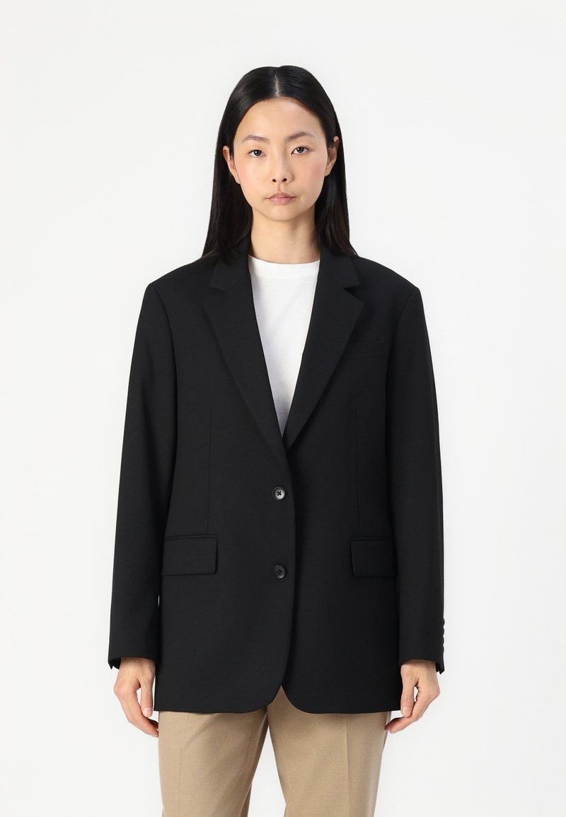 Schwarzer Blazer mit taillierter Passform, mit Reverskragen, zwei Knöpfen und vorderen Taschen. Getragen über einem weißen Hemd, aus glattem Stoff, mit strukturiertem Schnitt.