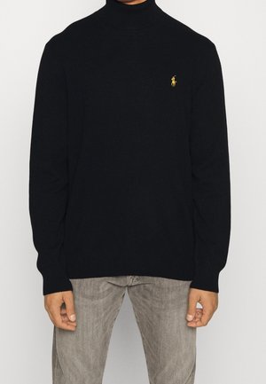 Mand iført en sort højhalset sweater med gul broderet logo på brystet og lysegrå bukser, stående mod en hvid baggrund.