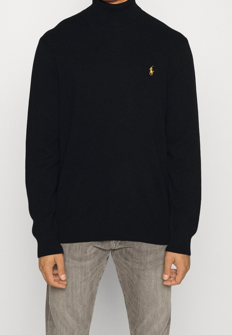 Homme portant un pull à col roulé noir avec un logo jaune brodé sur la poitrine et un pantalon gris clair, debout devant un fond blanc.