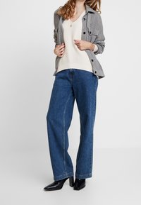 Chaqueta de camisa a cuadros gris sobre un suéter de punto crema y jeans de pierna ancha azules. Botines negros con tacón completan el conjunto.