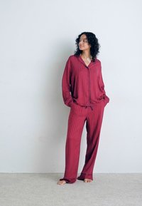 Marinrandig pyjamasset i mjukt tyg; knäppbar topp med krage och långa ärmar, parat med löst sittande byxor. Barfotamodell står.