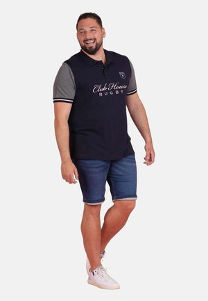 Homme souriant portant un polo noir et gris "Club House Rugby", un short en jean et des baskets blanches, debout sur un fond blanc.