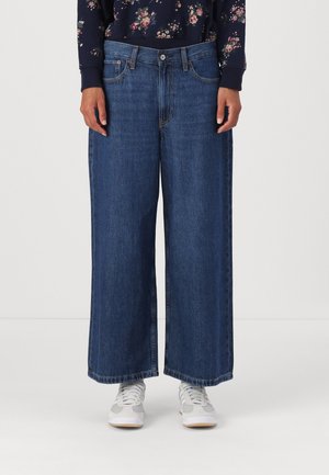 Wijd gesneden donkerblauwe denim jeans met een hoge taille, met voorzakken en een rauwe zoom. Gepaard met lichtgrijze sneakers.