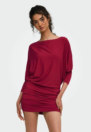 Jeune femme aux cheveux bouclés portant une robe rouge cintrée avec de longues manches chauve-souris sur un fond clair uni.