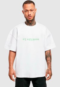 Bílé oversized tričko s zeleným grafickým textem "21 STUDIOS EST MMX" uprostřed hrudníku. Bavlněný materiál, neformální design, krátké rukávy.