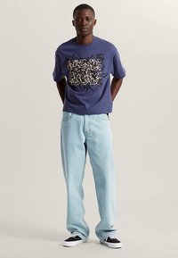 Maglietta di cotone blu navy con design grafico astratto beige e nero, abbinata a jeans di denim azzurro chiaro e scarpe da ginnastica nere.