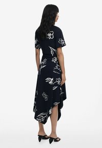 Schwarzes Kleid mit weißem Blumenmuster, kurzen Ärmeln, asymmetrischem Saum, weichem Stoff und fließendem Design. Getragen mit schwarzen Sandalen.