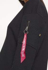 Sweat-shirt noir avec poche zippée sur la manche et étiquette en tissu rouge fixée indiquant "REMOVE BEFORE FLIGHT". Texture lisse, design décontracté.