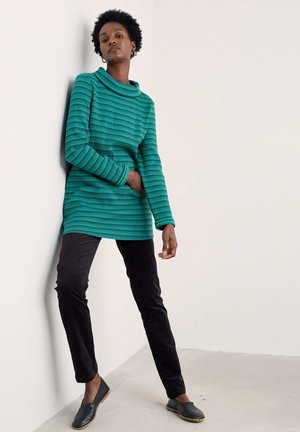 Türkis gestreiftes Strick-Tunika mit hohem Kragen, kombiniert mit schwarzen Slim-Pants und dunklen Slippern. Einfaches, strukturiertes Design.