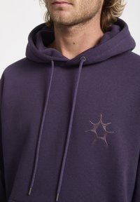 Purple hoodie made of soft fabric, featuring an embroidered star-like logo on the chest, with contrasting drawstrings and metal toggles.
