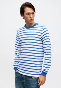 Mężczyzna ubrany w niebiesko-biały koszulę w poziome paski z długim rękawem, niebieskimi mankietami i kołnierzykiem, w zestawieniu z niebieskimi dżinsami, stoi na białym tle.