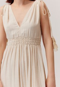 Beżowa sukienka z dekoltem w serek, marszczonym pasem i miękkim, teksturowanym materiałem. Posiada plecione ramiączka z frędzlami na ramionach.