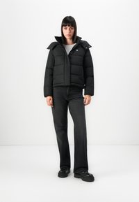 Femme se tenant contre un fond blanc, portant une doudoune noire, un haut blanc, un jean noir et des chaussures à plateforme noires.